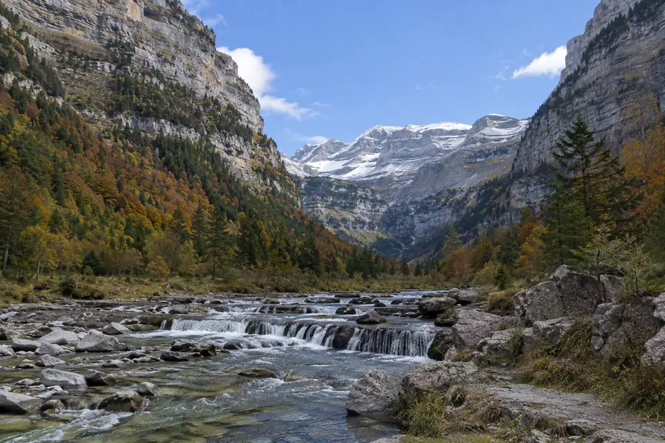 Breed dal met steile gelaagde kalkwanden, herfstkleurig bos en een ondiepe rivier met watervalletjes.