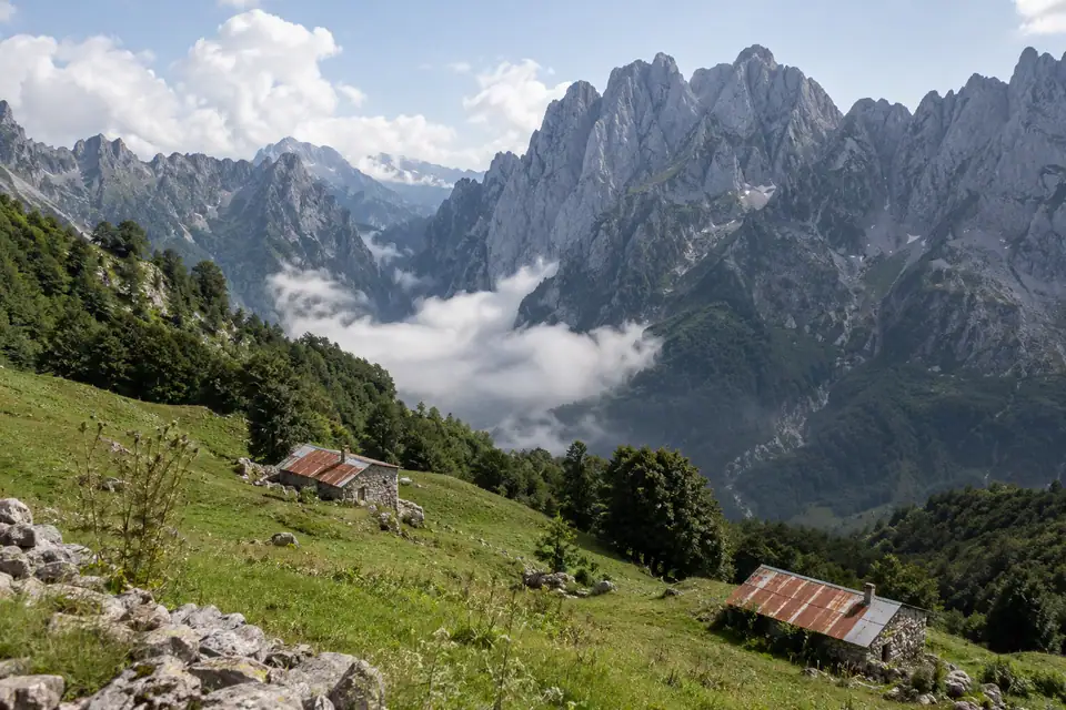 Steile grijze kalkstenen toppen boven felgroene weiden, met donkere beukenbossen en lage stenen hutten.