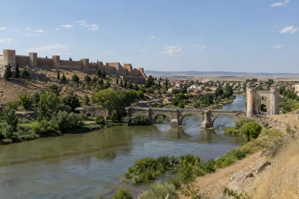 Roodbruine vestingmuren met vierkante torens boven een rivier met een lange lage stenen brug.