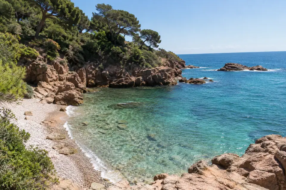 Kleine kiezelbaaitjes tussen roodachtige rotspunten, met parasolpijnen en struiken tot aan het heldere turquoise water.