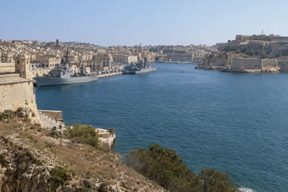 Grijze marineschepen liggen langs de kades van een diepe haven, met oude forten op kale heuvels.