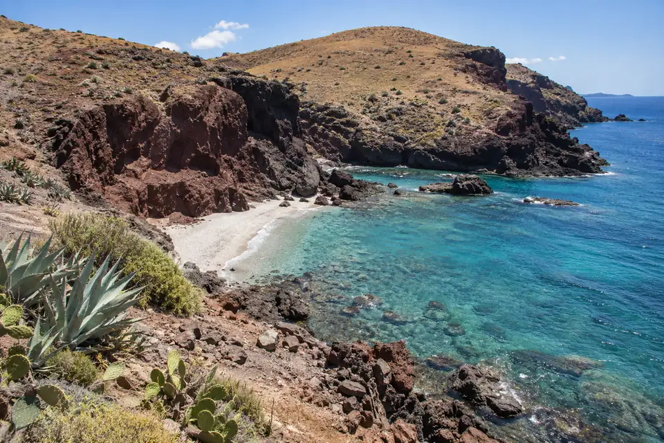 Roodbruine vulkaankliffen boven turquoise water met agaves en cactussen op de kale hellingen.