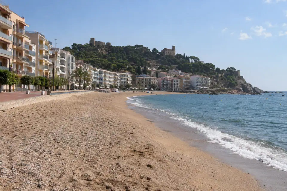 Een lang recht strand met betegelde boulevard en flats, en een beboste heuvel met kasteelmuur erachter.