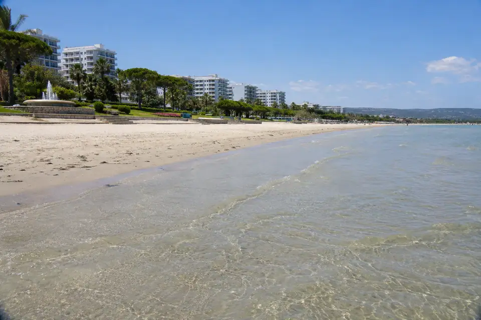 Een zeer breed vlak strand met fijn licht zand, met gazons, pijnbomen en witte hotelblokken erachter.