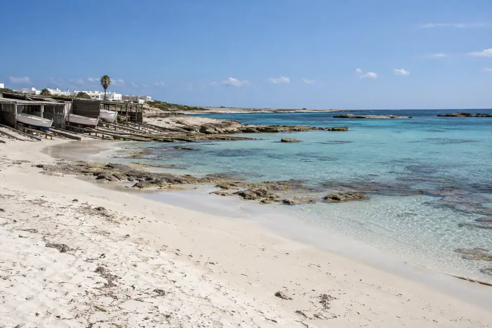 Breed bijna wit strand met lage rotsplaten, houten botenhuisjes en ondiep aquamarijn water.