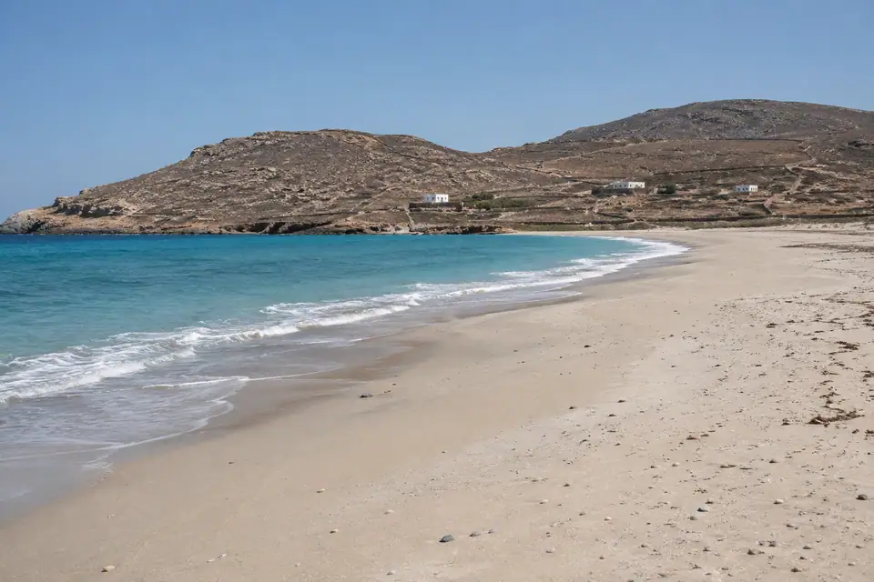 Breed leeg strand van bleek zand langs turkoois water, met kale bruine heuvels erachter.