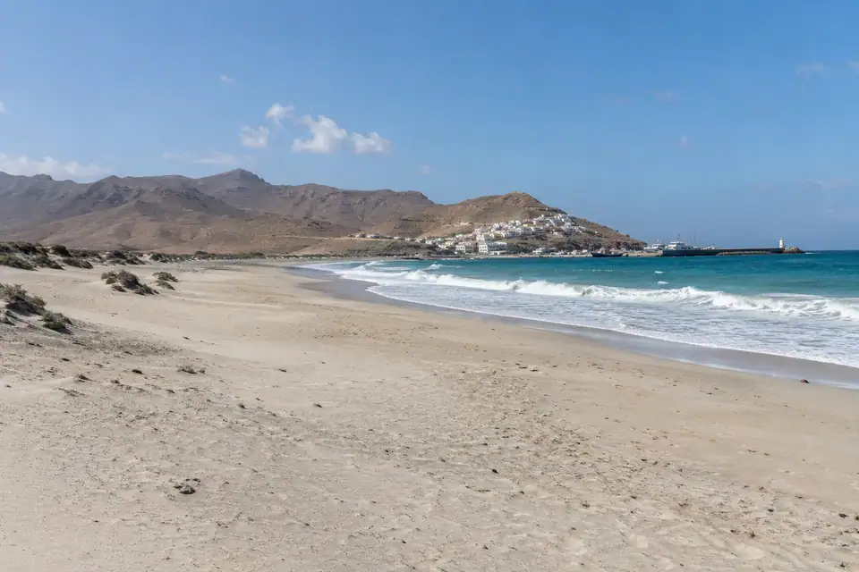 Zeer breed bleek strand met laag duin, kale bruine bergen en een compacte witte stad.