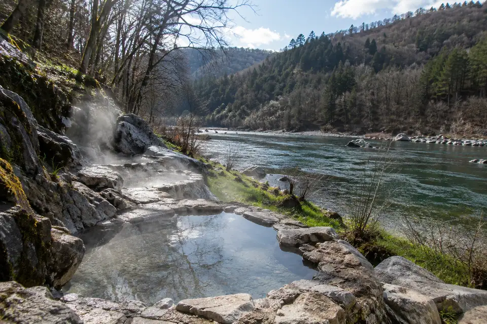 Ondiepe stenen badjes met dampend warm water in een groene oever langs een brede rivier.
