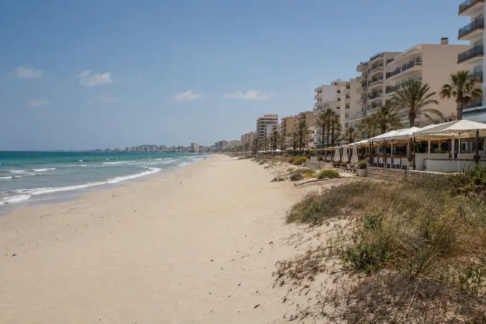 Vlakke lange strook bleek zand met een aaneengesloten rij hotelblokken en strandbars onder witte luifels.
