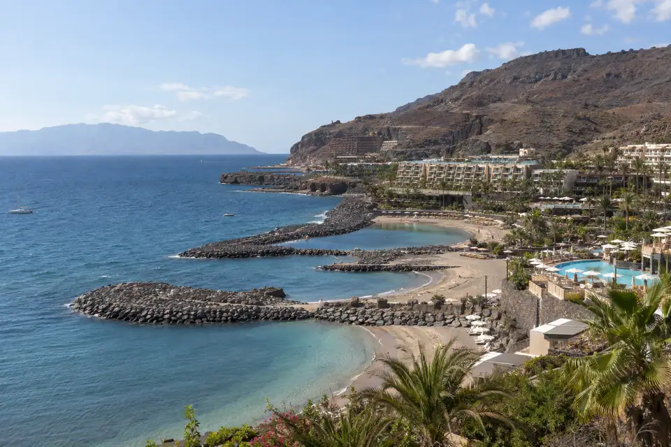 Grote hotelcomplexen met palmtuinen en zwembaden boven kleine strandjes tussen golfbrekers, erachter droge bruine heuvels.