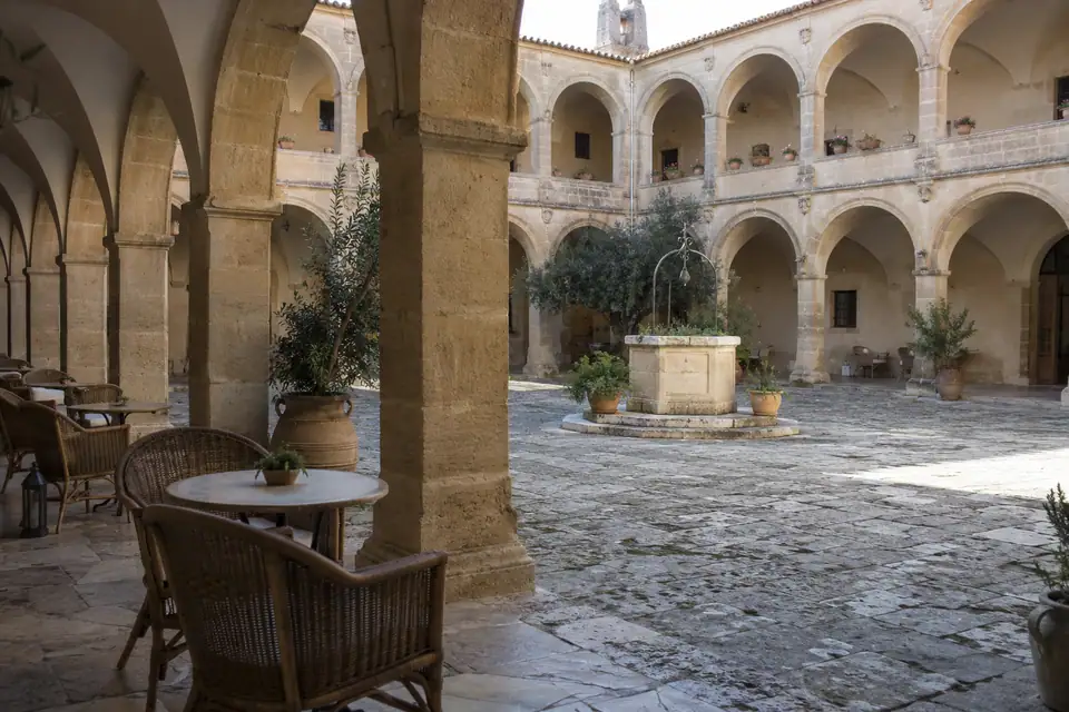 Kloosterhof met twee verdiepingen rondbooggalerijen rond een stenen put, met rieten stoelen onder de arcade.