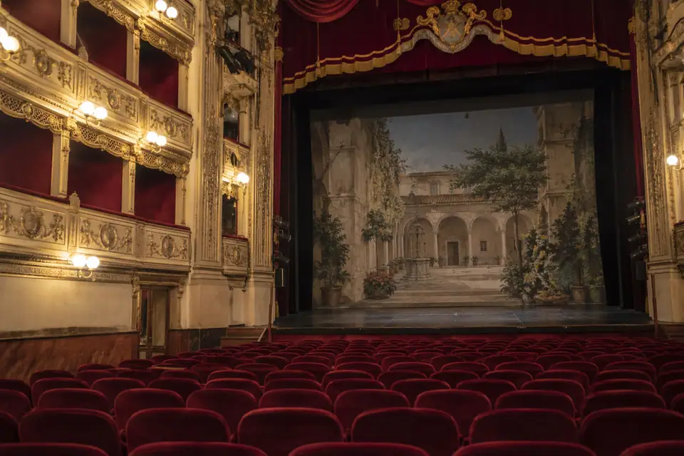 Oud theater met rode fluwelen stoelen, vergulde balkons en een beschilderd decor op het podium.