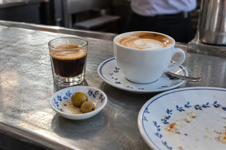 Klein glaasje koffie naast een grote kop, en een schaaltje olijven bij een groot bord.