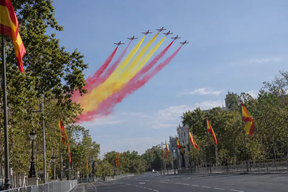 Vliegtuigen in formatie trekken rode en gele rookpluimen boven een brede laan met vlaggenmasten.