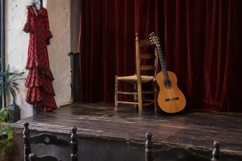 Een houten stoel met gitaar op een klein podium, daarachter een zwaar donkerrood gordijn.