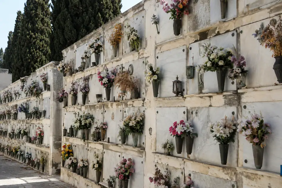 Hoge kerkhofmuur met vijf lagen grafnissen achter marmeren platen en vazen met kunstbloemen.