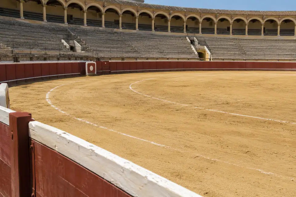 Lege ronde arena met geel zand, een rood houten hek en betonnen tribunes erboven.