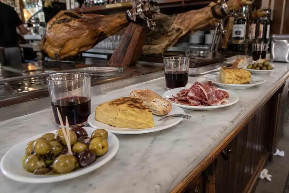 Marmeren bar vol kleine witte bordjes met olijven, aardappelomelet en ham, daarachter hangende hammen.