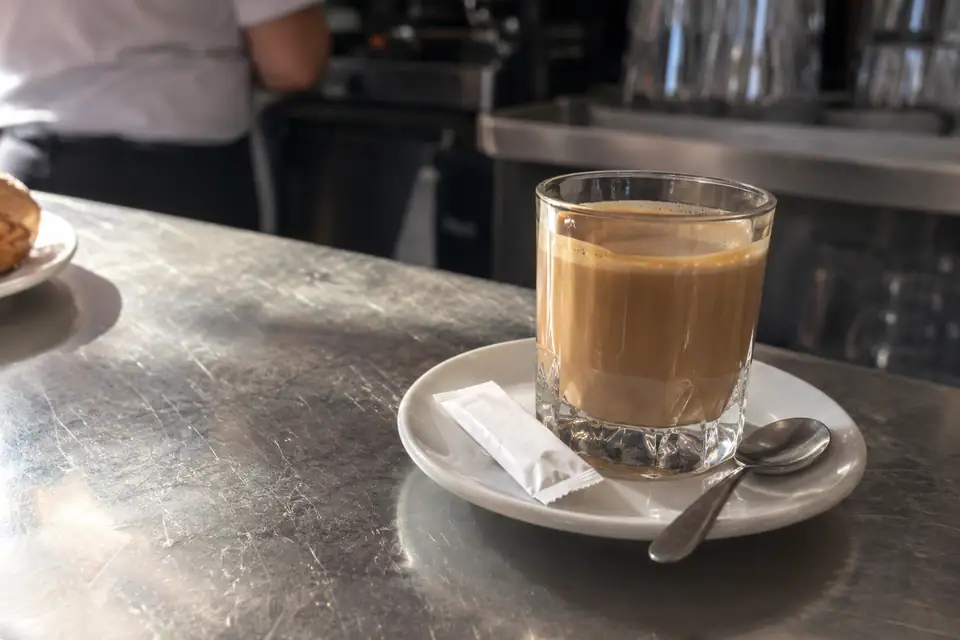Een dik glas lichtbruine koffie met melk op een schoteltje met een zakje suiker.