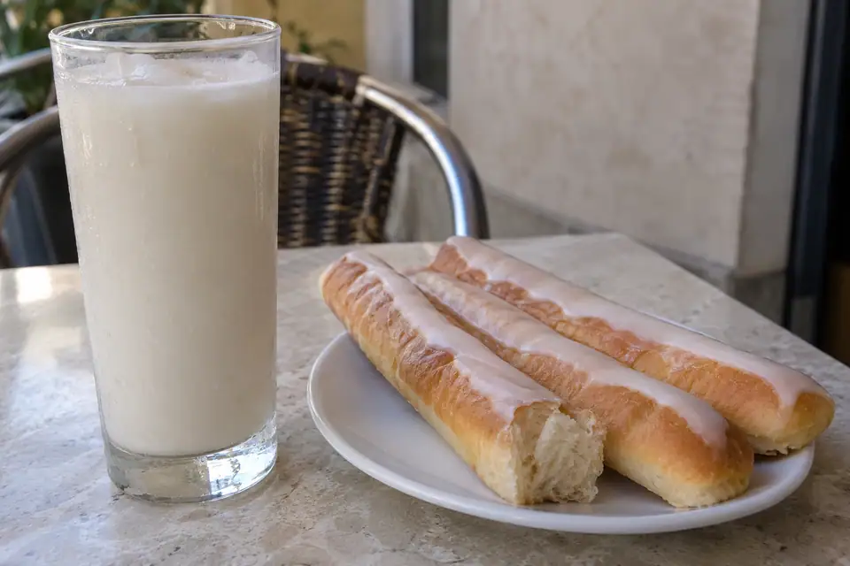 Een hoog beslagen glas met een melkwitte drank, naast een bord langwerpige geglazuurde koekjes.