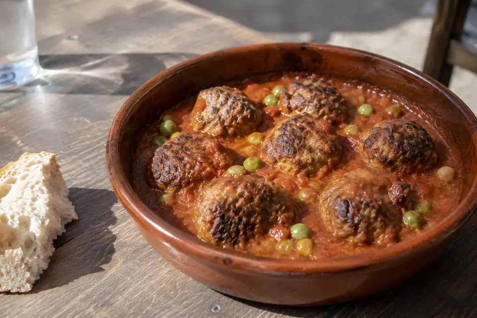 Acht gehaktballetjes half in dikke roodbruine tomatensaus in een aardewerken schaal, met erwtjes ertussen.