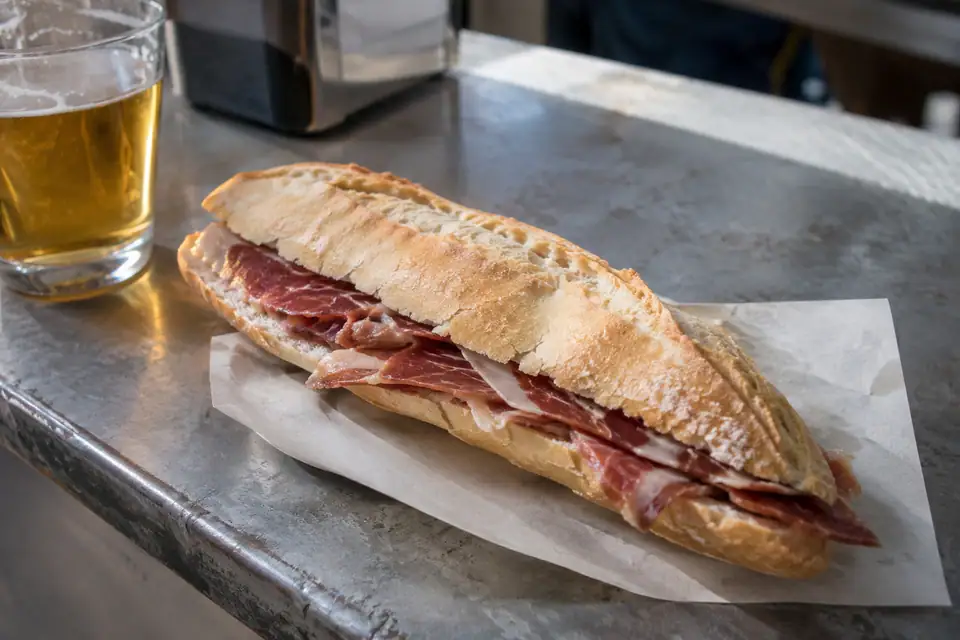 Een lang krokant stokbrood met dunne sneetjes donkerrode ham, op papier op een zinken bar.