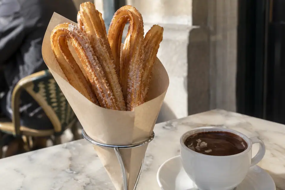 Lange geribbelde gefrituurde deegstengels met suiker in een papieren puntzak naast dikke donkere chocolademelk.