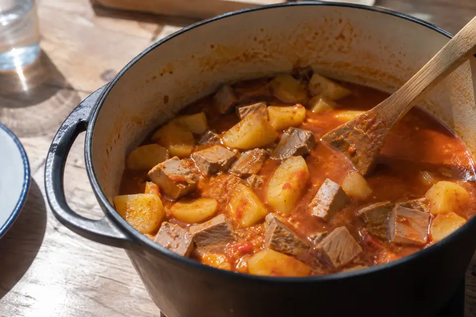 Diepe pan met grove stukken aardappel en donkere tonijn in oranjerode paprikabouillon, houten pollepel erin.