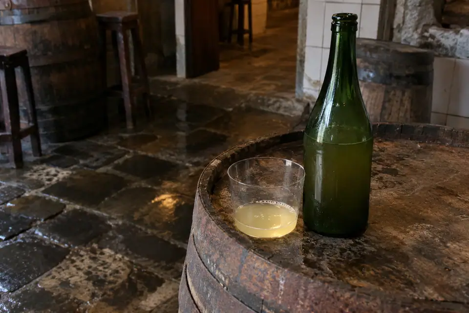 Donkergroene fles troebele cider naast een breed glas met een bodempje, op een vat.