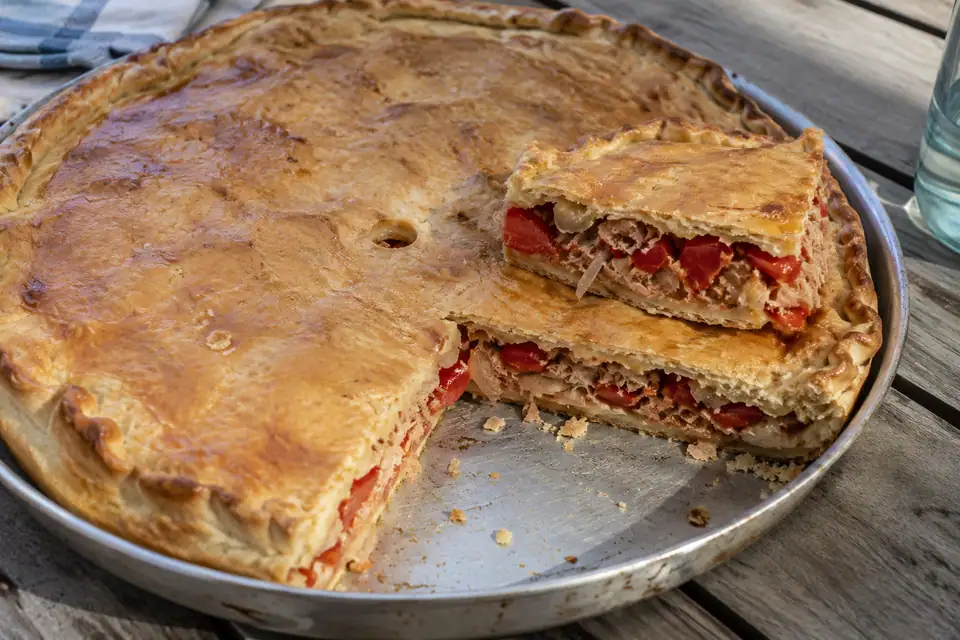Platte ronde deegtaart met goudbruine korst, twee stukken uitgesneden met vulling van tonijn en paprika.