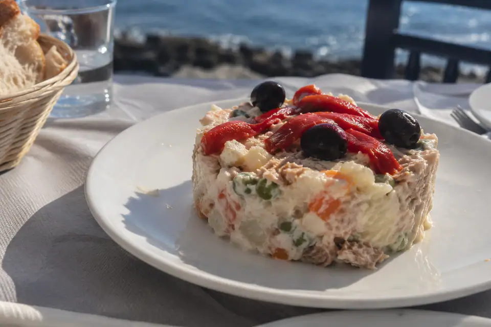 Romige aardappelsalade met worteltjes en erwtjes op een wit bord, met paprikareepjes en zwarte olijven.