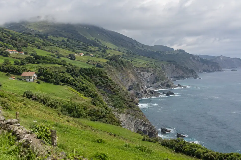 Steile groene heuvels lopen naar een grijsblauwe baai, met lage wolken op de toppen.