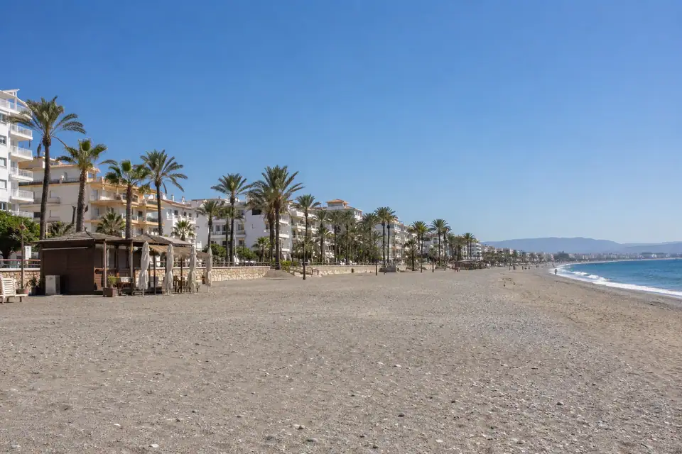 Lang grijsgoud strand met witte appartementen langs een palmenboulevard, steile grijze bergrug erachter.