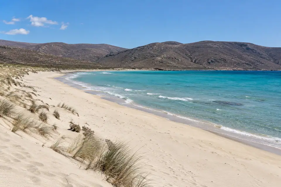 Zeer lang licht zandstrand met ondiep turquoise water en kale bruine heuvels zonder bomen.